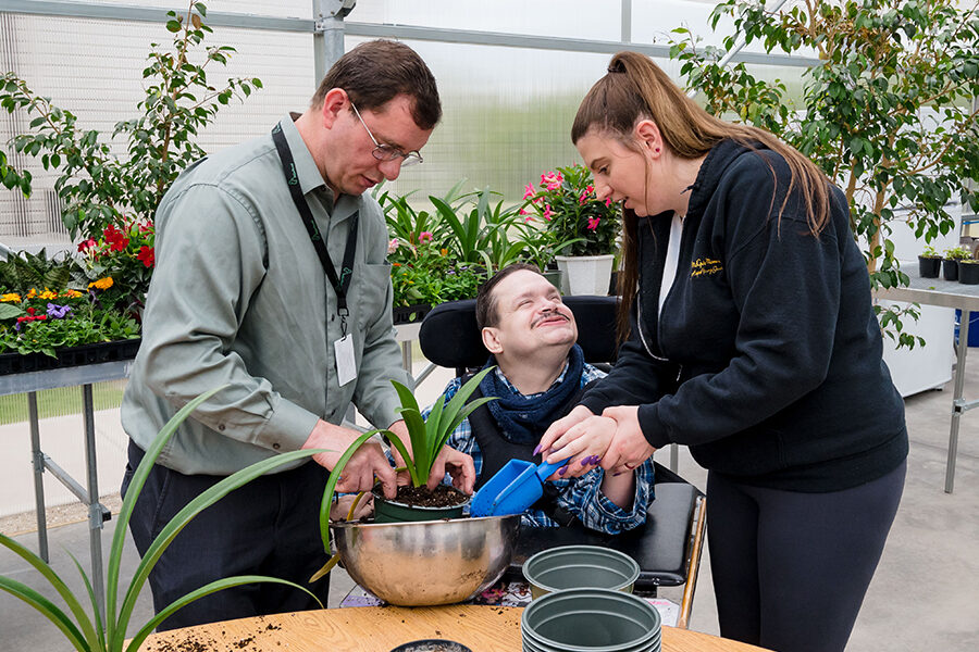 Two McGuire Memorial team members helping a gentlman in a wheelchair pot a plant int he McGuire Memorial greenhouse.