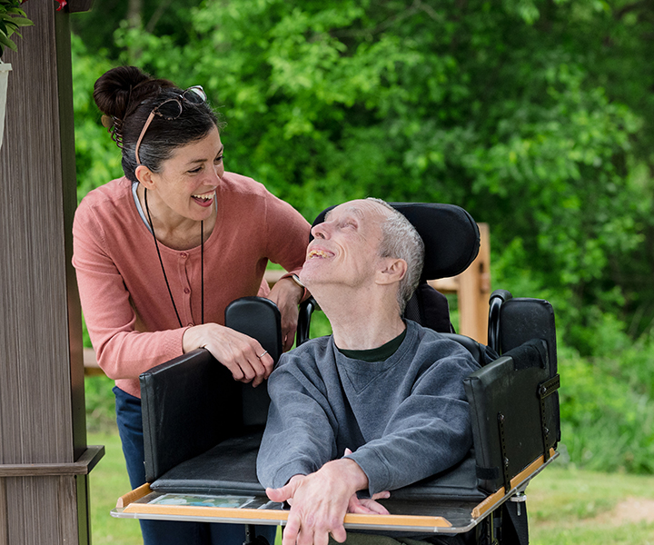 Smiling resident in a wheelchair on the St. Francis Nature Trail. Resident is smiling at a McGuire team member who is stnading next to him.