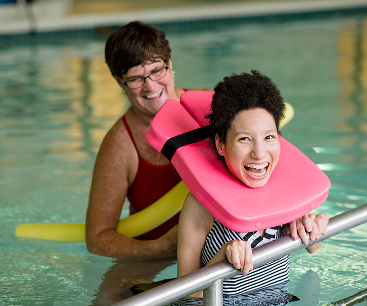 Smiling resident in the swimming pool wearing a pink floatation device. Also in the pool is a smiling instructor