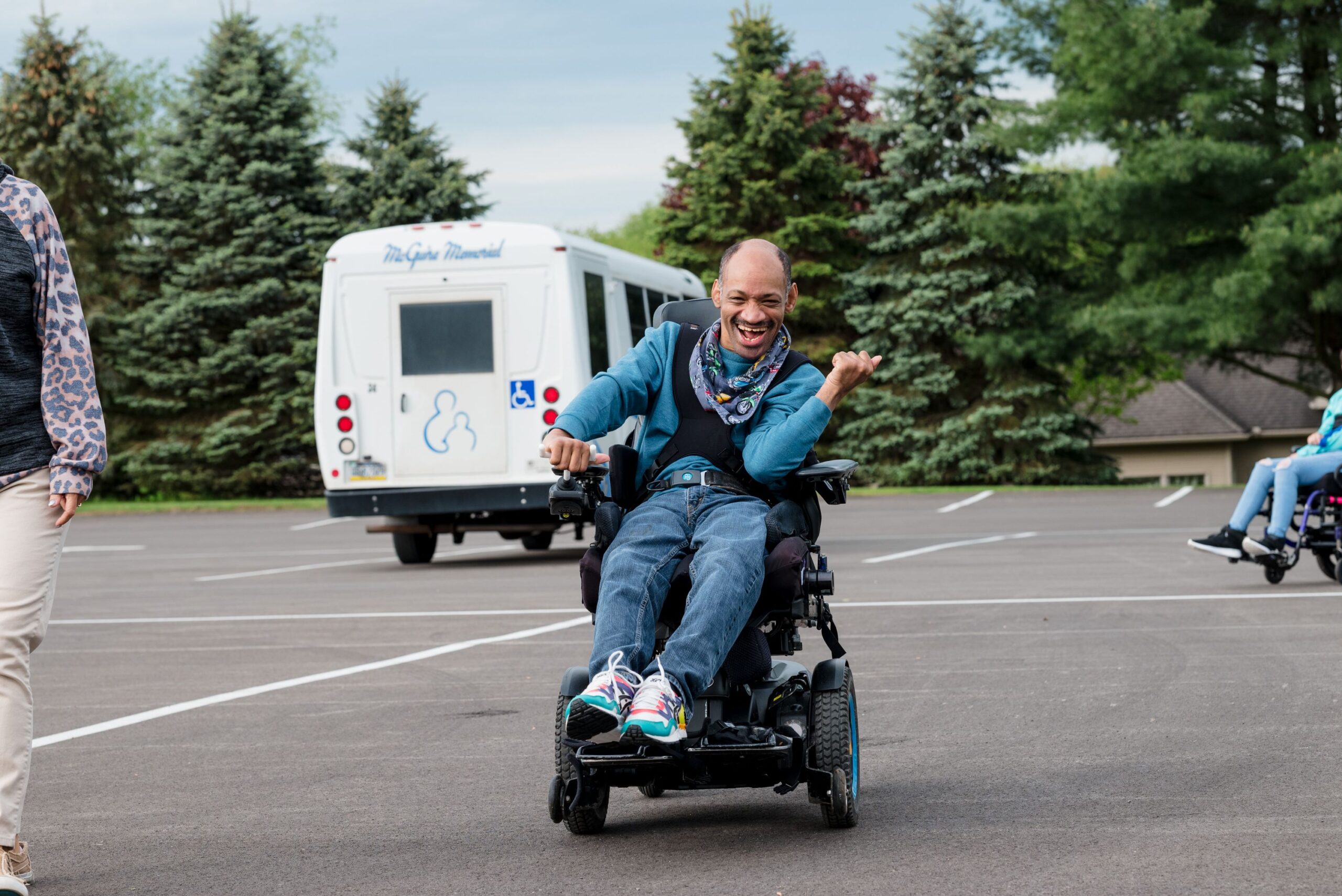 A smiling gentleman in a wheelchair. He is driving his chair through a parking lot. In the background is a wheelchair accessible van. 