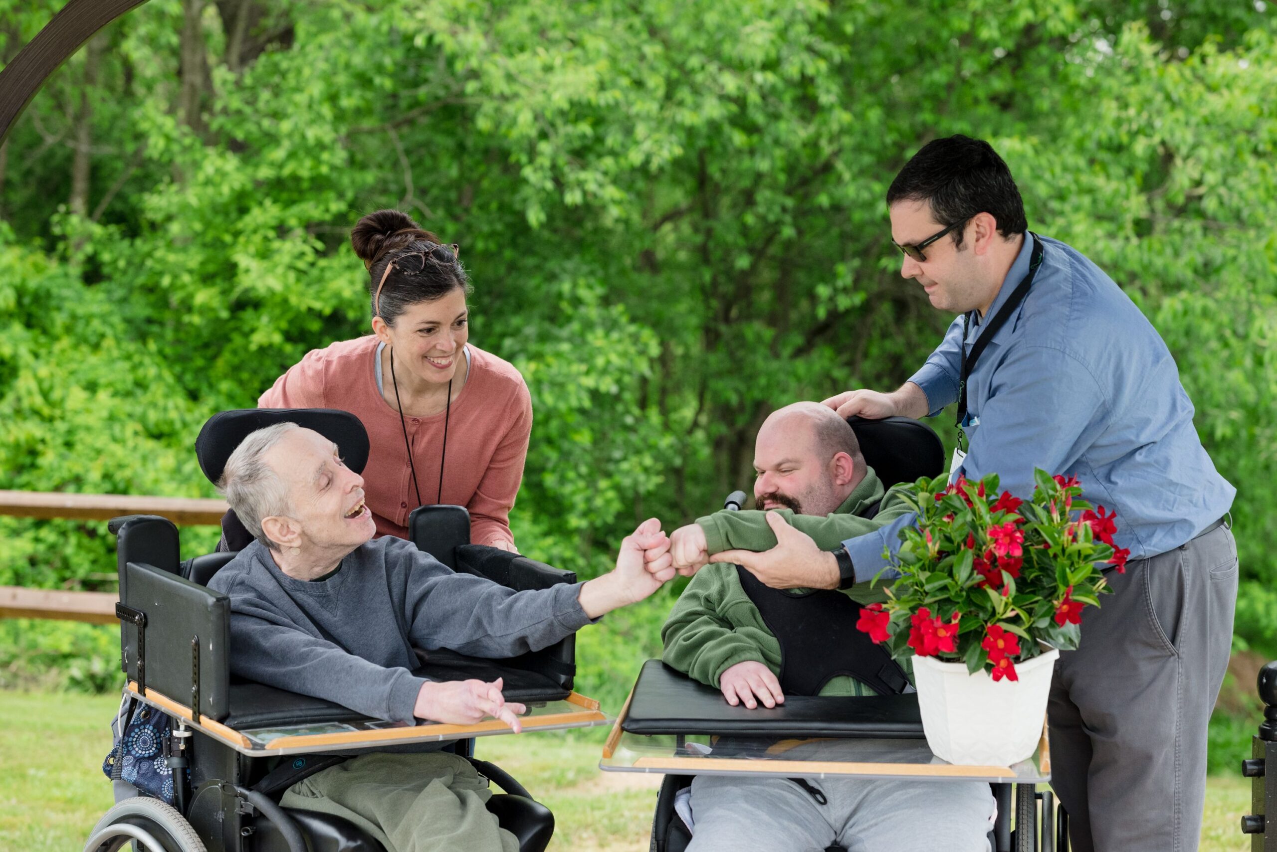 Two residents in wheel chairs outside on the St. Francis Nature Trail. The residents are giving each other a fist pump. Also in the picture are two McGuire Memorial team members helping them. 