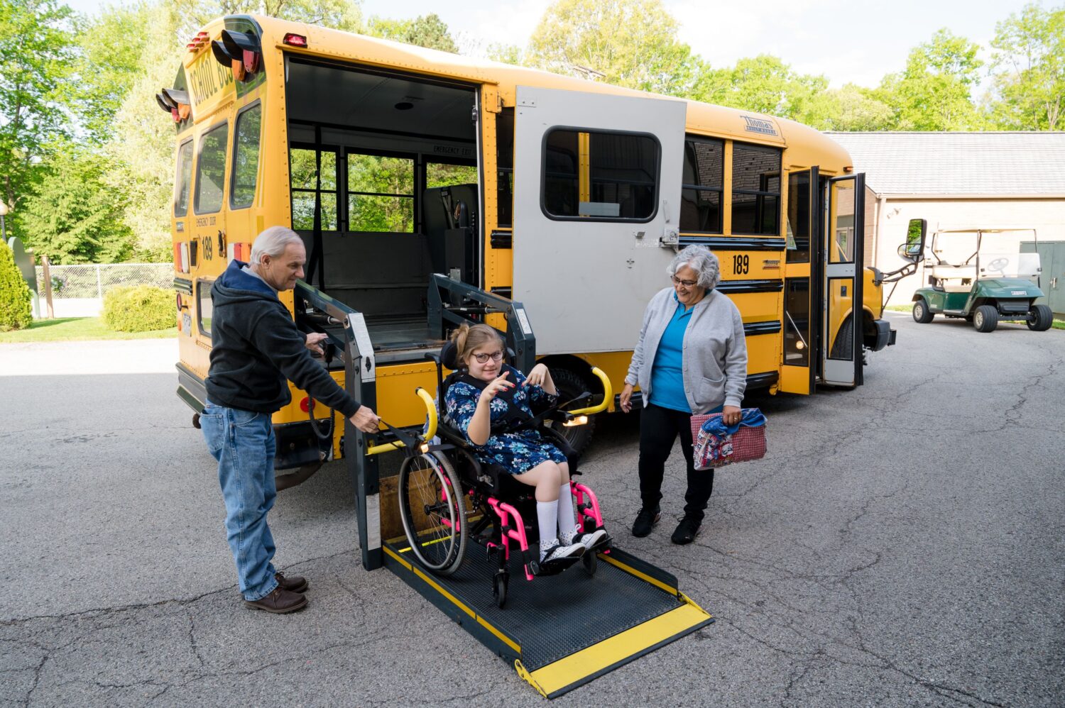 A young girl in a wheelchair is being helped off of a school bus by two McGuire Memorial team members