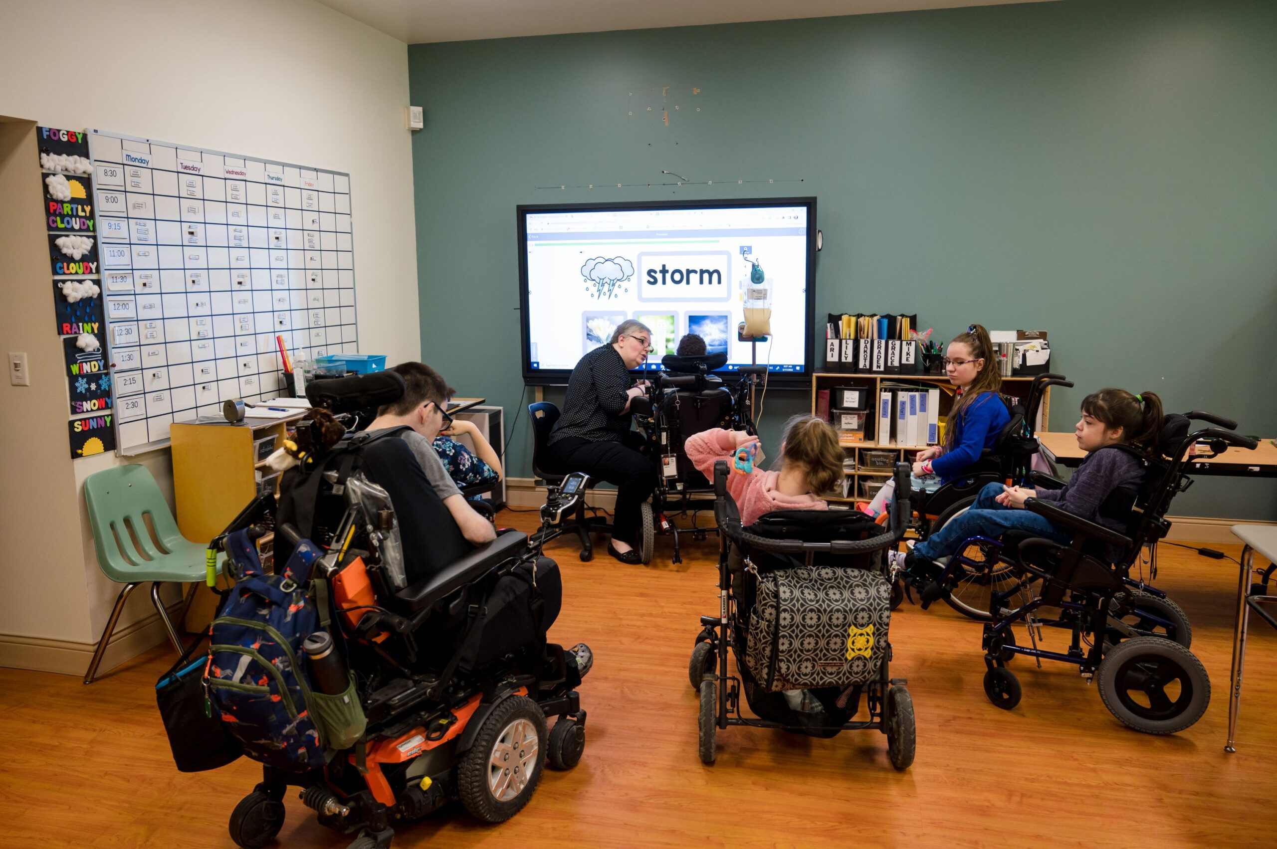Classroom setting with six students in wheelchairs and a teacher. The teacher is helping a student in front of a screen with pictures and words on it. 