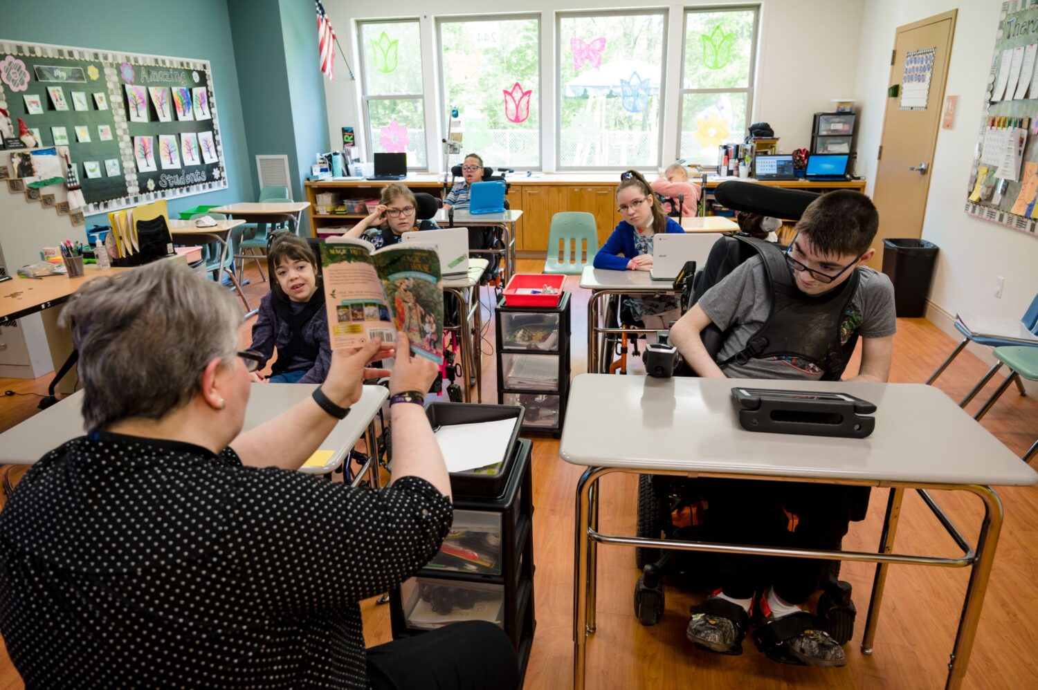 A teacher reading to students. The students are in wheelchairs and sitting at desks while she reads to them. 