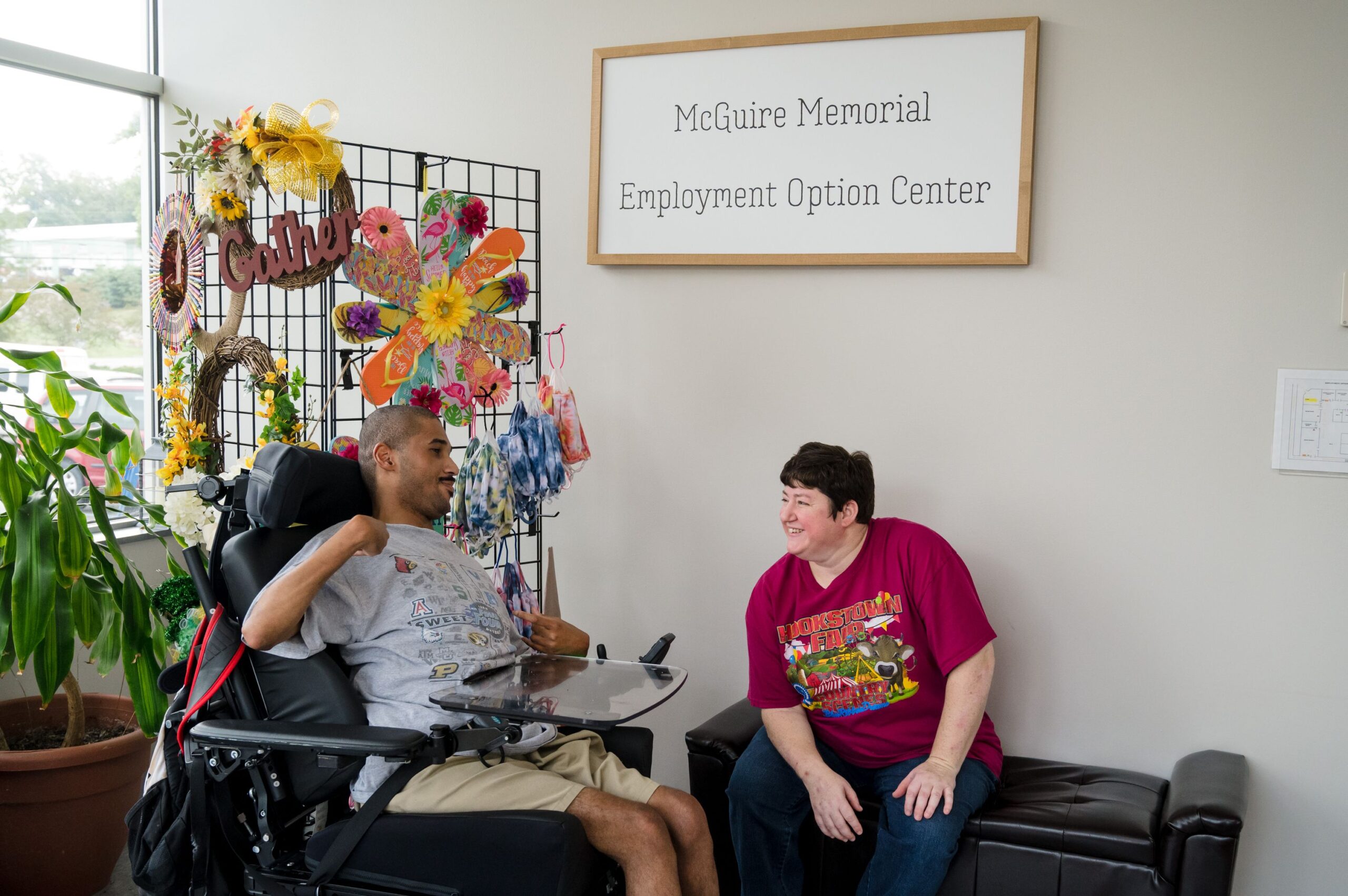 A gentleman in a wheelchair talking to a McGuire Memorial team member. Behind him is a rack holding colorful signs and wreaths 