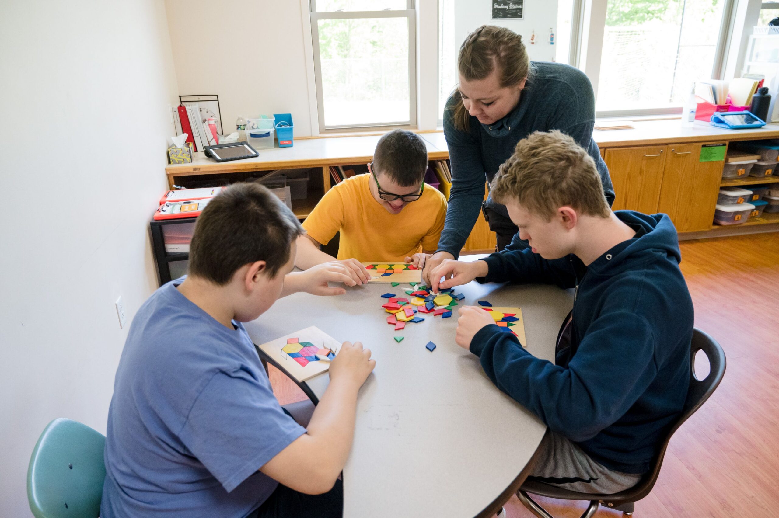 Three older students sitting at a desk each working on a puzzle. Helping them is a teacher. 