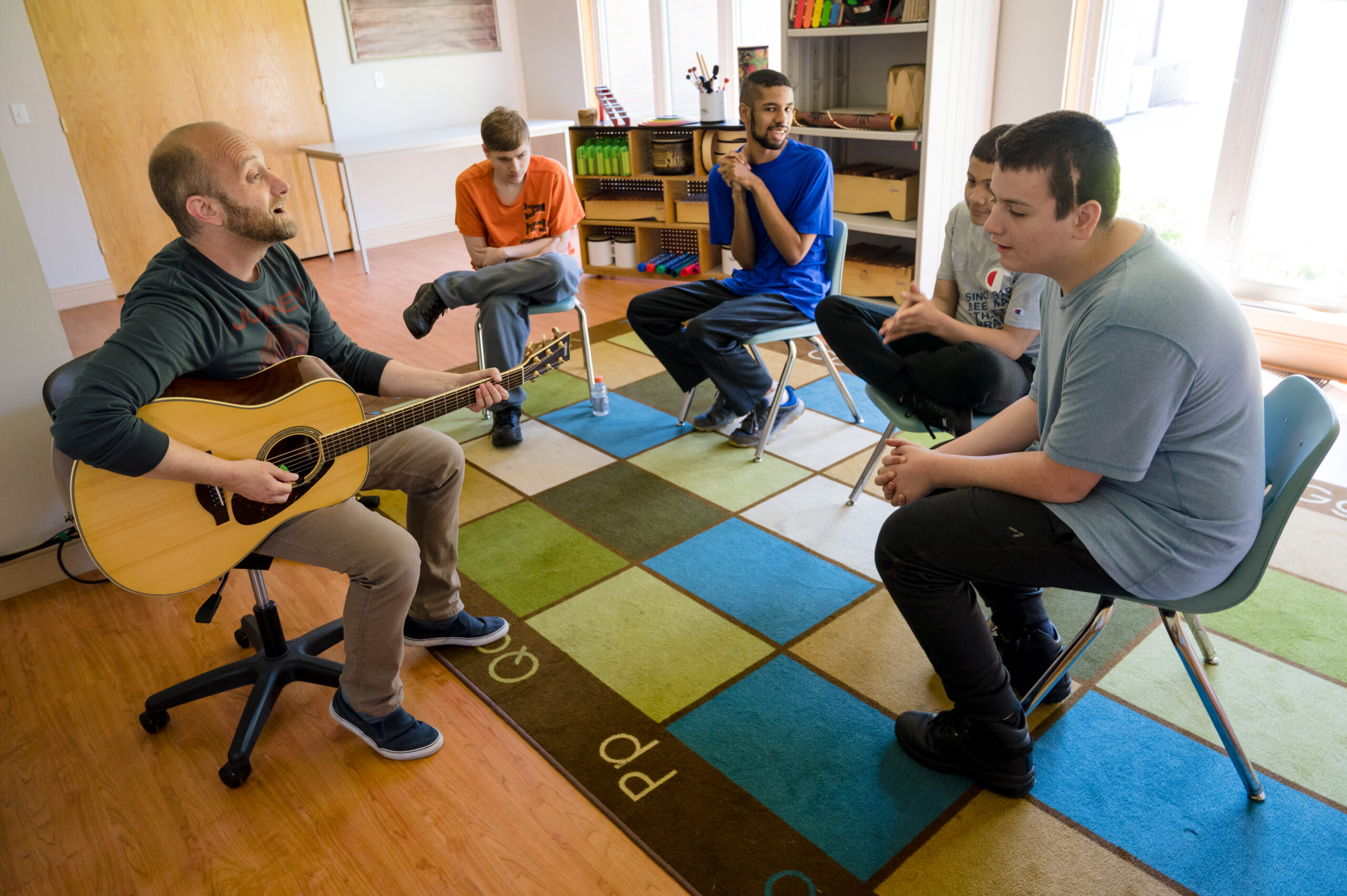 A music teacher with a group of teenagers sitting in a circle. The teacher is playing a guitar. 