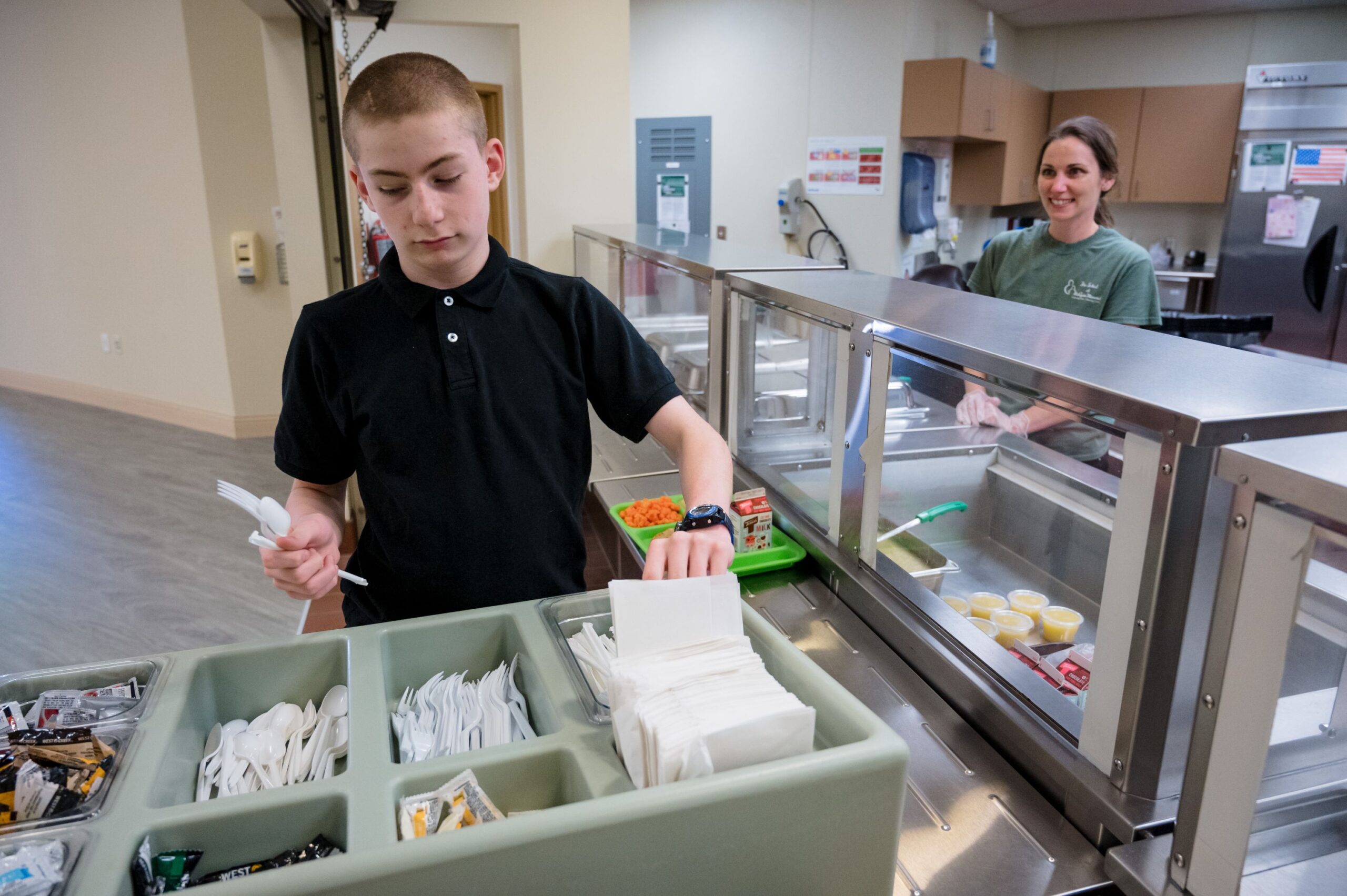 A student in the cafeteria getting silverware to eat lunch. Behind him is a McGuire Memorial team member who served him lunch. 