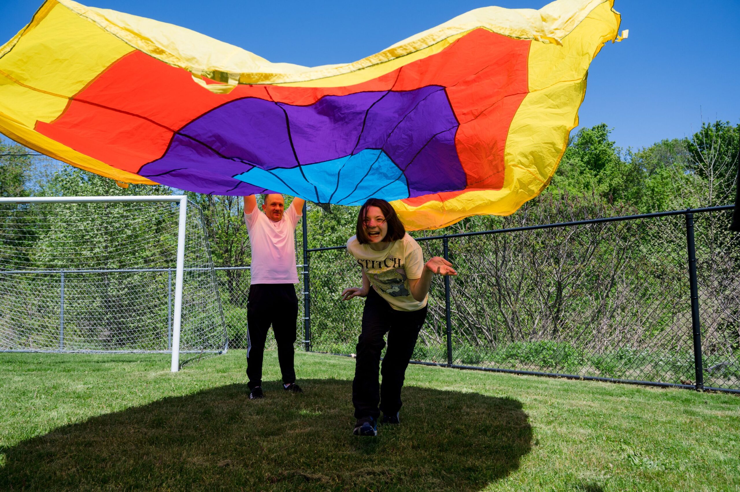 A female student is running under a rainbow colored parachute that a teacher is lifting in the air. 
