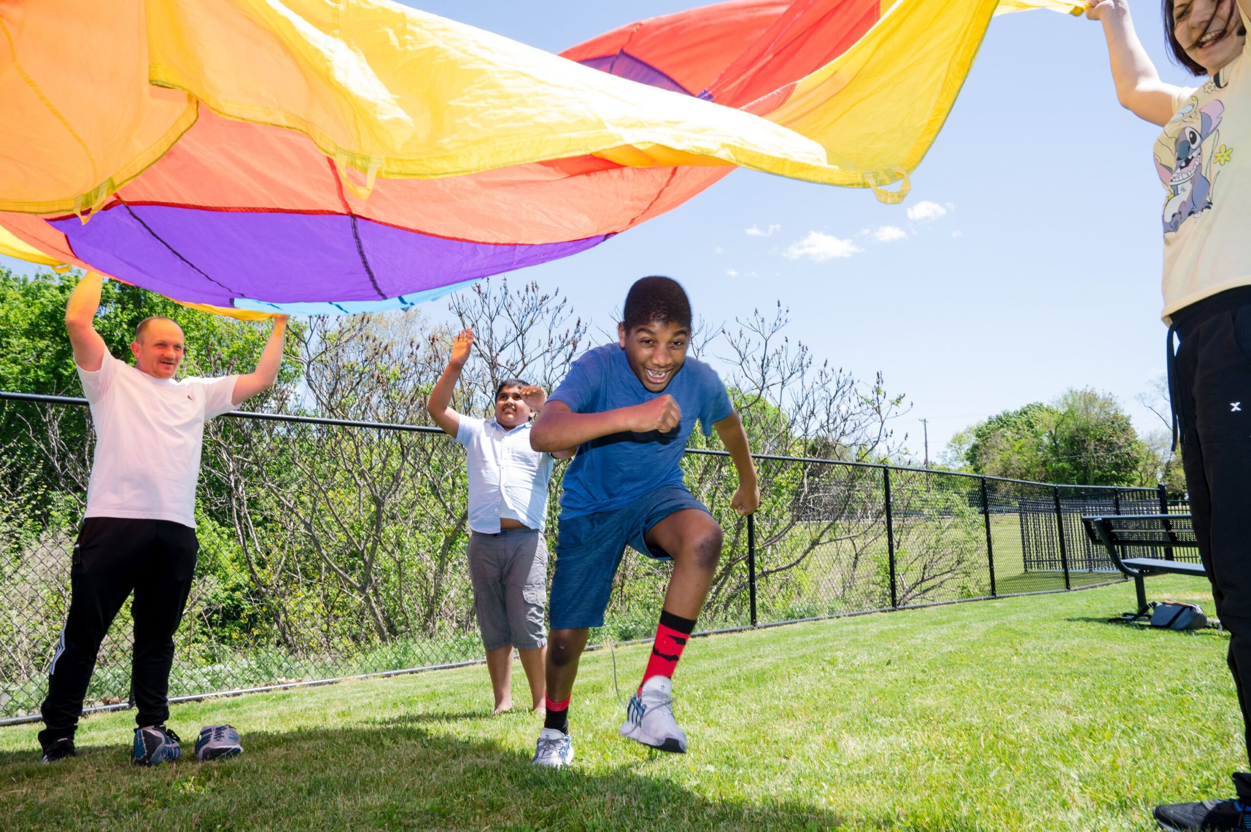 A student running outside underneath of a rainbow colored tent. A teacher and another student are lifting the tent in the air.  
